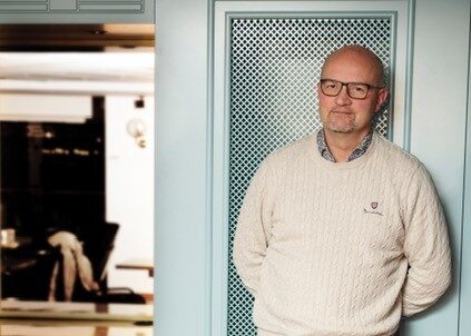 Mederålders man med glasögon som lutar mot en ljustblå vägg